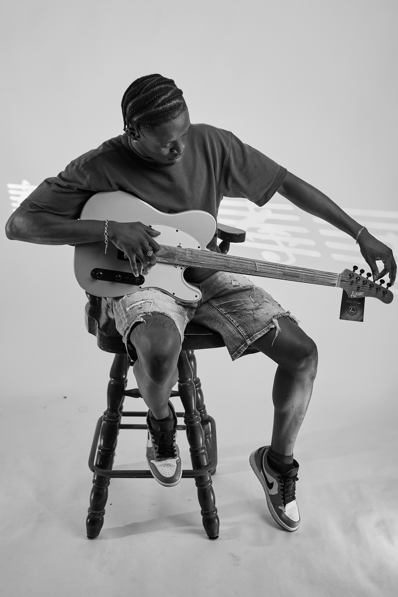 Black-and-white portrait of Ceddy seated on a stool, tuning a bass guitar.