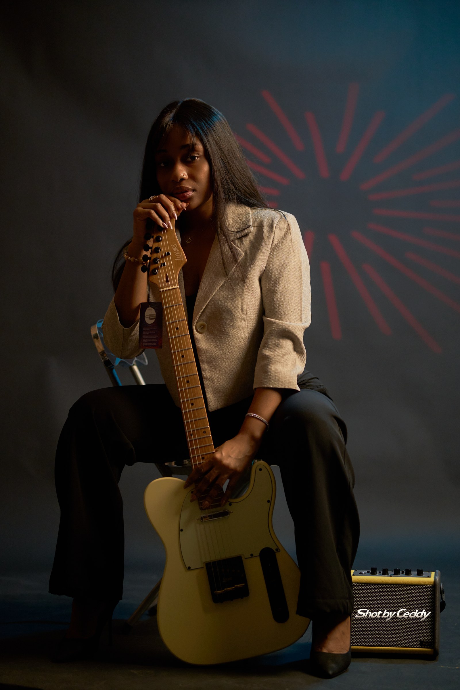 Full seated portrait with a cream electric guitar, gold jacket, and red patterned background.