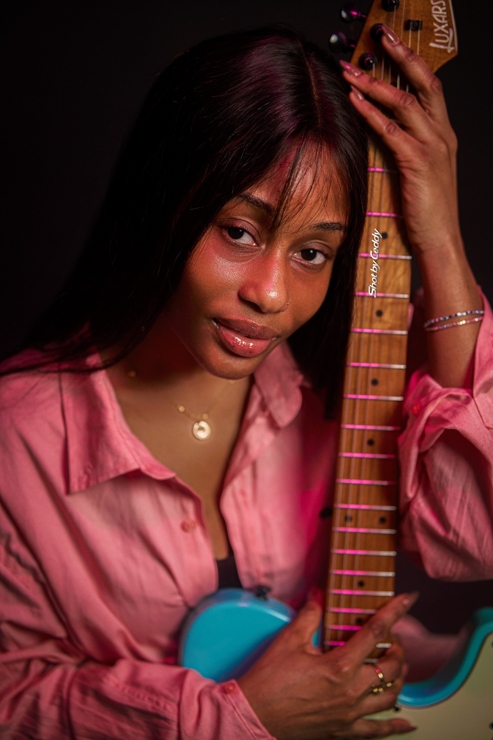 Close portrait in a pink satin shirt beside the neck of a turquoise electric guitar.