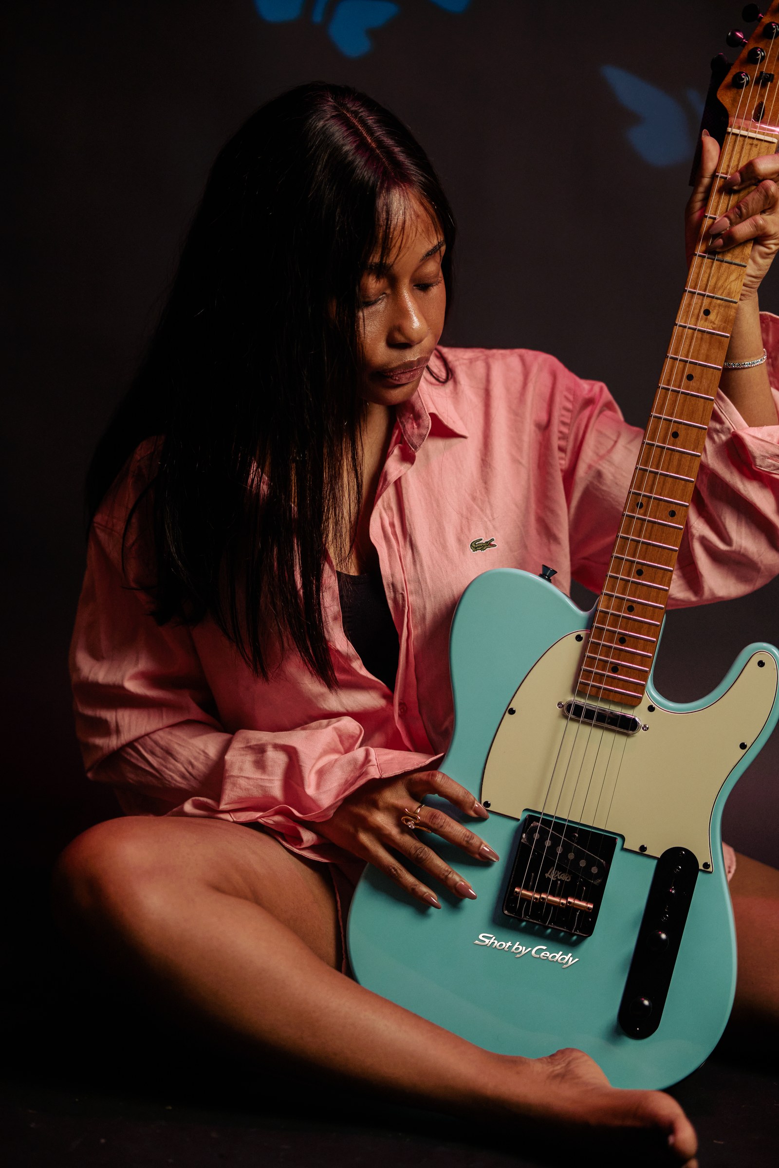 Seated portrait looking toward a turquoise electric guitar, wearing a pink satin shirt.