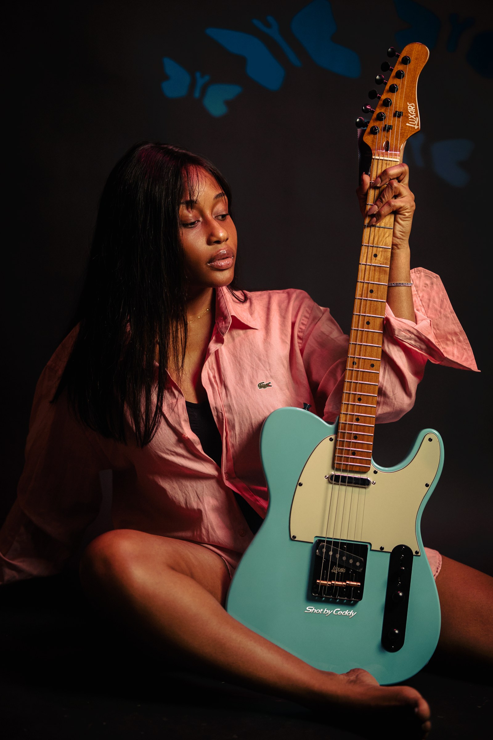 Portrait holding a turquoise electric guitar upright against a blue patterned backdrop.