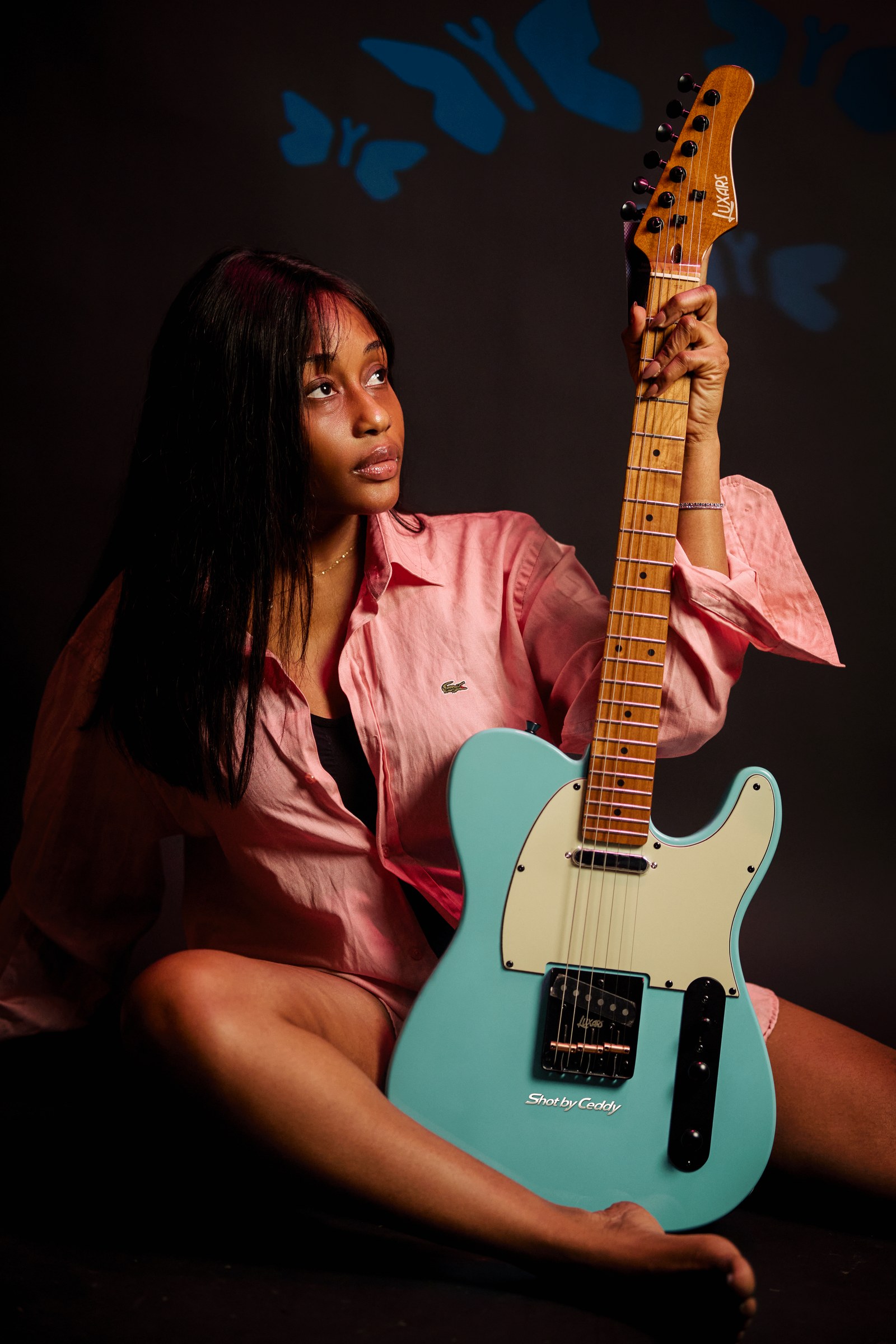 Seated portrait in pink satin with a turquoise guitar and blue studio patterns.