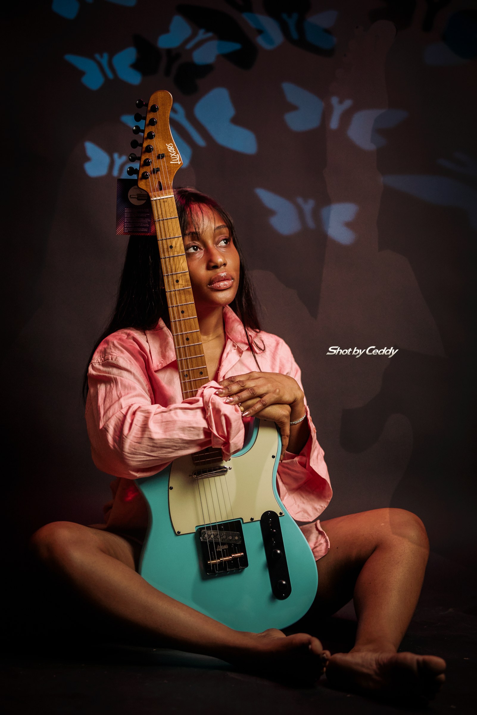 Wide seated pose holding a turquoise guitar vertically, wearing a pink satin shirt.