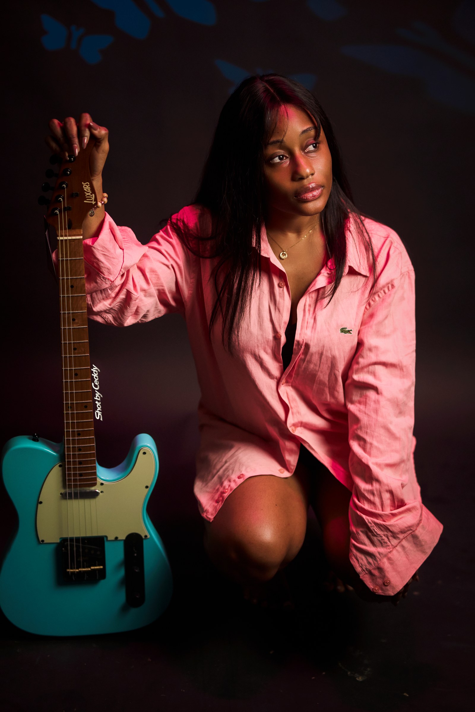 Seated portrait in pink satin, one hand lifted beside a turquoise electric guitar.